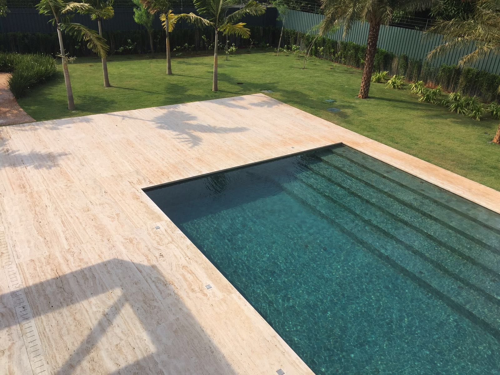Vista de cima de uma piscina retangular com deck largo de travertino romano claro e o tanque revestido em pedra escura, com degraus ocupando toda a largura