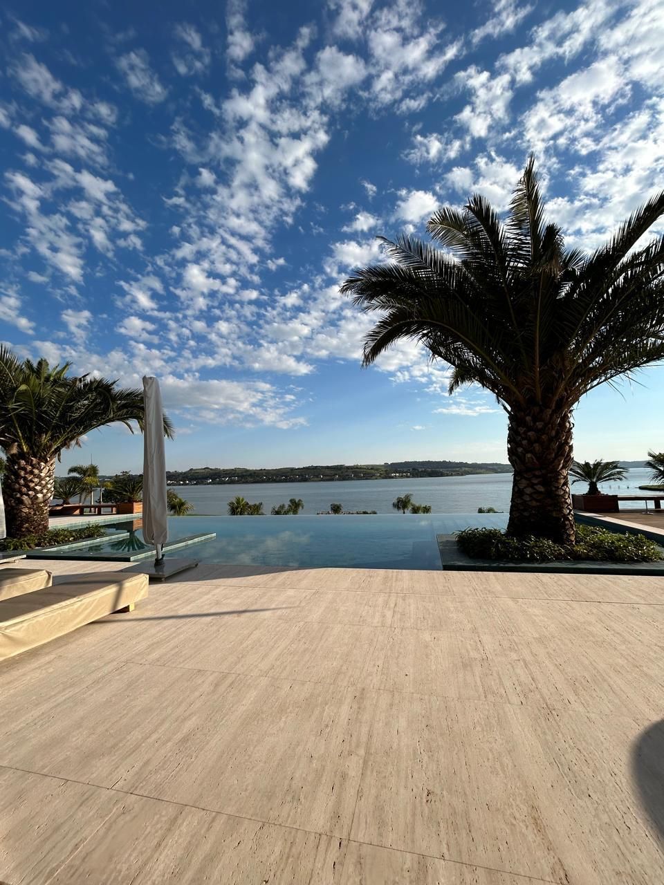 Deck externo em ladrilho de travertino romano light, com palmeiras e vista aberta ao fundo