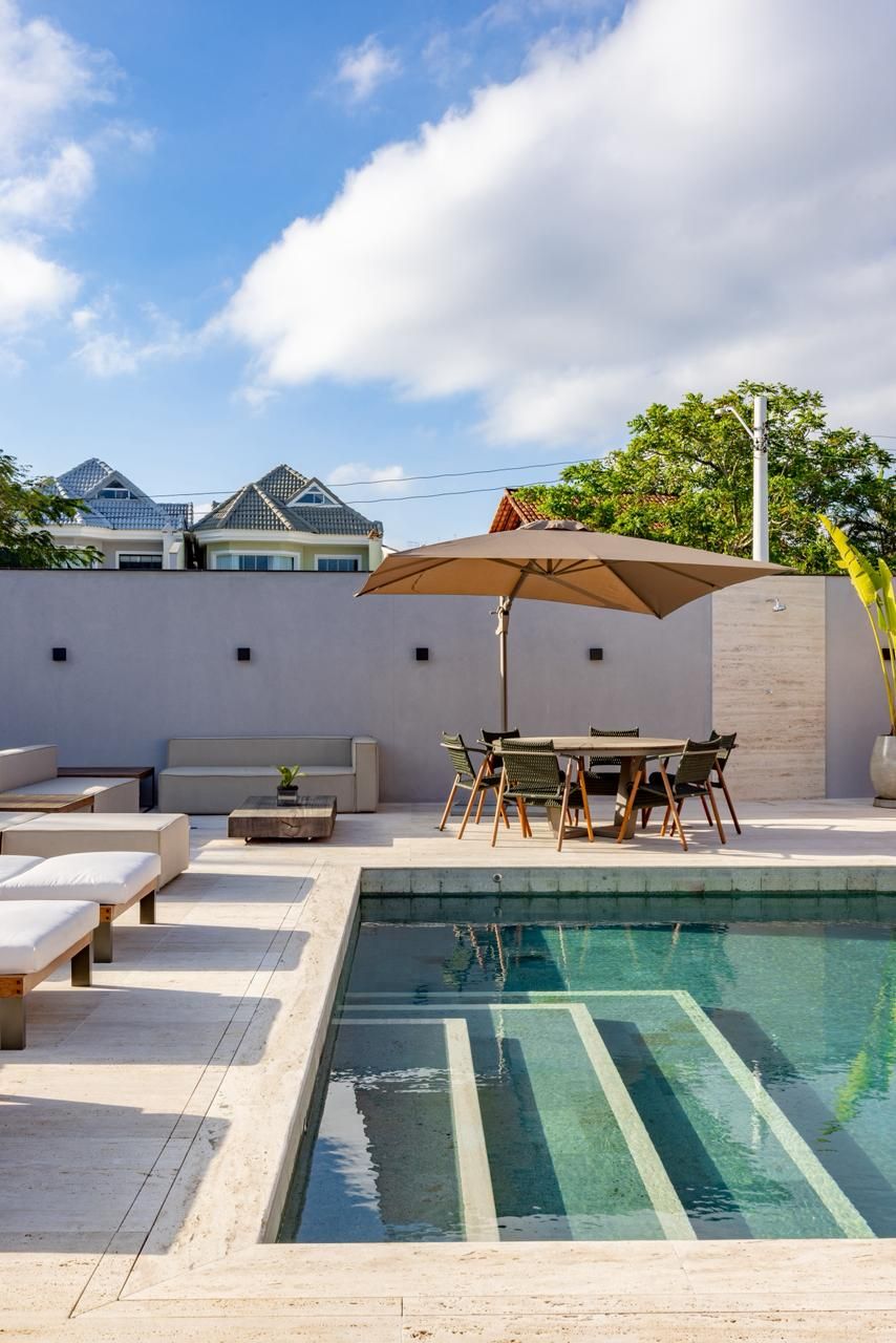 Piscina com deck de travertino romano light claro, guarda-sol, mesa de jantar externa e espreguiçadeiras