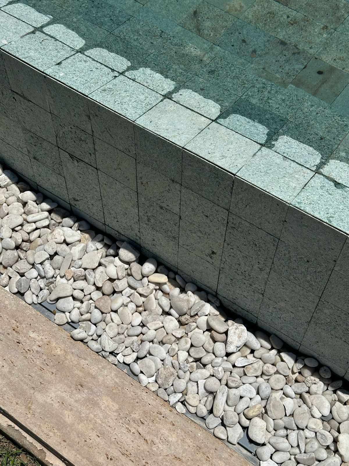 Detalhe da borda de piscina em Pedra Hijau Lisa, com o coroamento em peças verdes, a face externa em 10x10 e uma faixa de seixos brancos na base