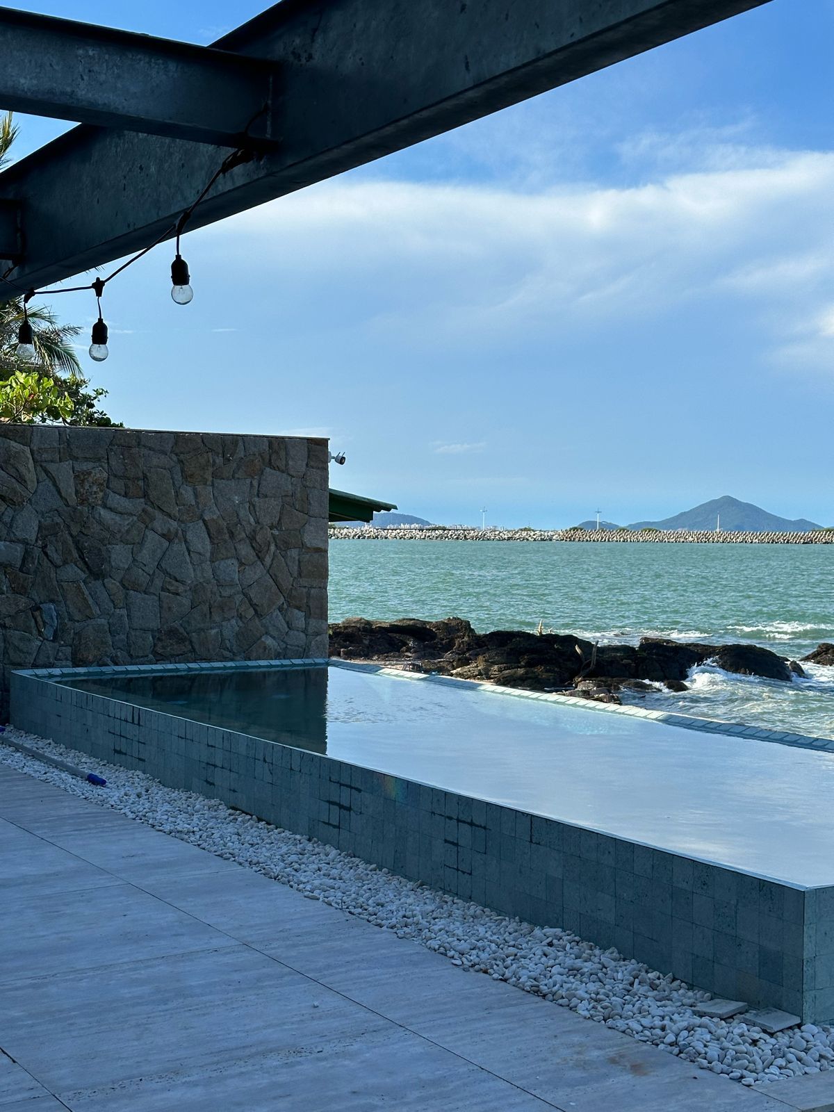 Piscina de borda infinita revestida em Pedra Hijau Lisa com a face externa à vista, muro de pedra natural ao lado e o mar logo atrás