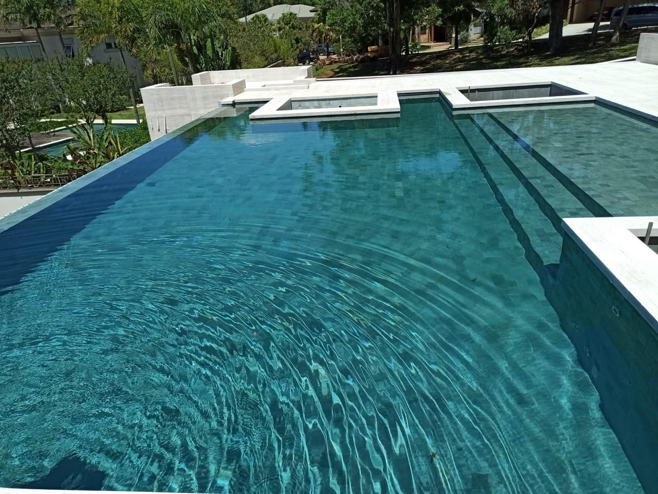 Piscina de borda infinita revestida em Pedra Hijau Lisa, vista de cima, com a água em verde turquesa, degraus largos de um lado e deck de travertino claro em volta