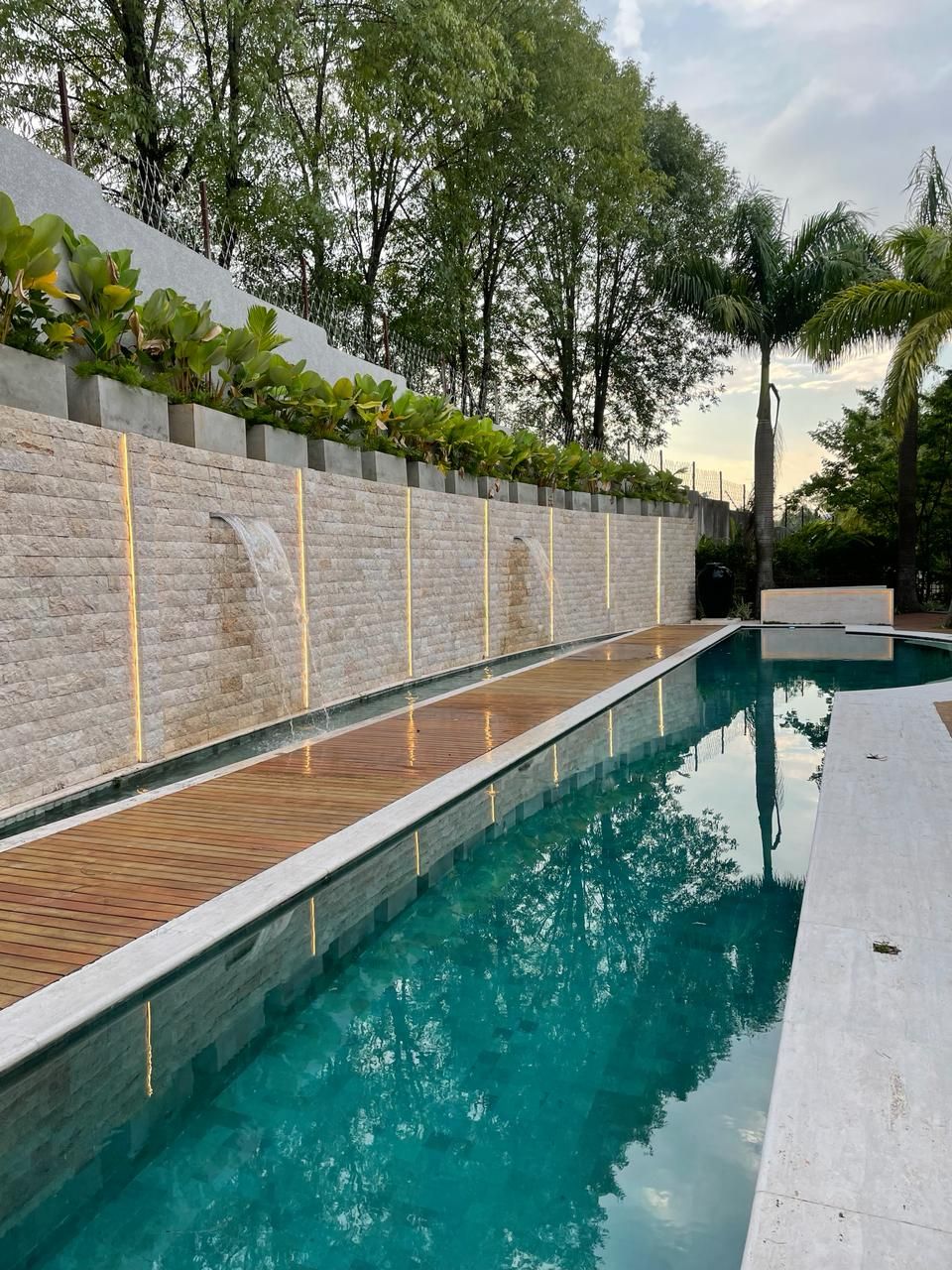 Muro comprido revestido em rock face de travertino romano ao lado de piscina de raia, com cascatas e iluminação linear