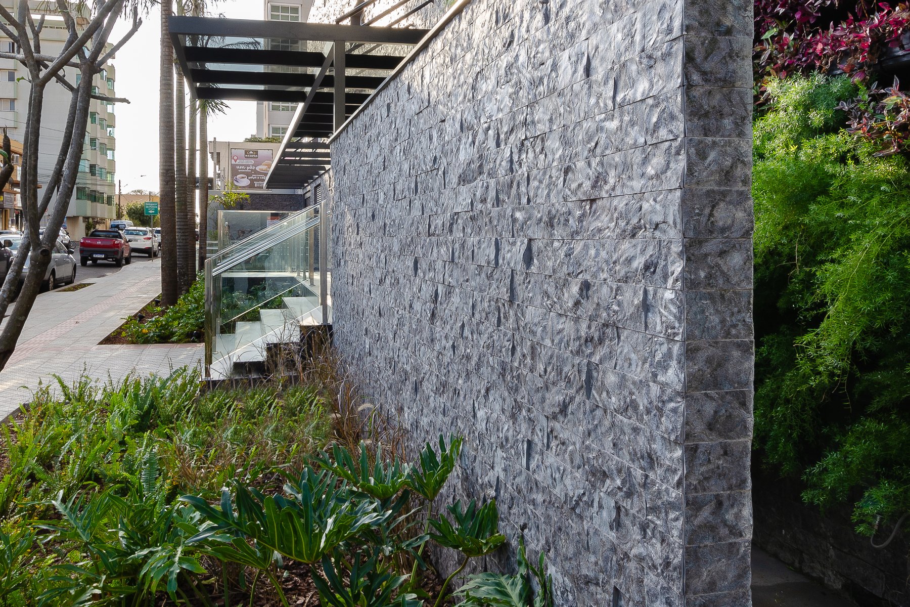 Muro alto revestido em rock face cinza, visto em perspectiva rasante, com jardim na base e prédios ao fundo