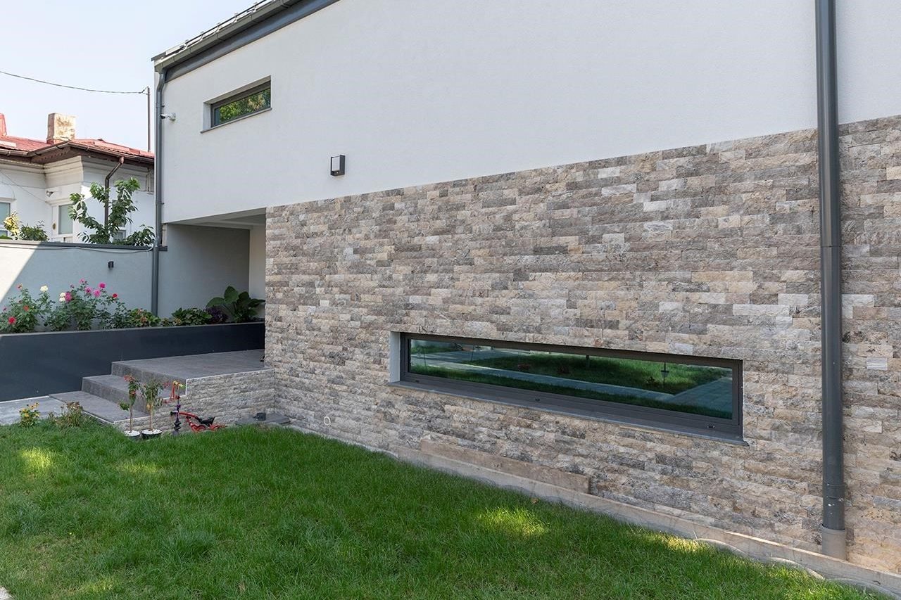 Parede externa de casa revestida em travertino rock face em tons de bege e cinza, com janela horizontal e gramado à frente